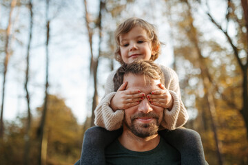 Wall Mural - Father walking with daughter on his shoulders across autumn forest, girl covering his eyes with palms. Dad and girl having fun outdoors.