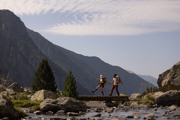 Two people are walking on a bridge over a river. The mountains in the background create a sense of awe and wonder. The scene is peaceful and serene, with the sound of the water