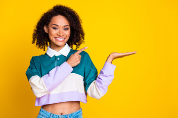 Wall Mural - Photo of cheerful woman with curly hair dressed striped top indicating at object on arm empty space isolated on yellow color background