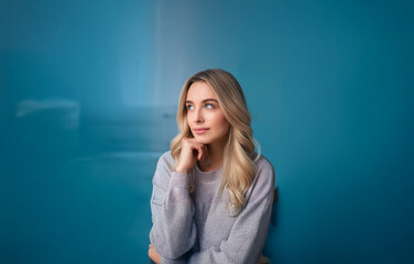 A young woman with long blonde hair and blue eyes is looking to her right, her hand resting on her chin.