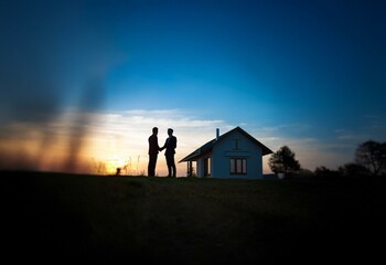 Two men in suits shake hands in front of a house at sunset.