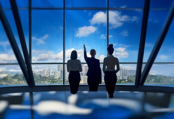 Three women in business attire stand in front of a large window overlooking a city skyline.