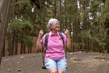 Happy senior woman sitting on swing in the forest enjoying relaxed and carefree moments, positive retirement lifestyle for attractive senior lady in pink shirt