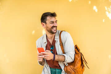 Young fashionable hipster freelancer businessman posing outdoor with yellow wall background and smart phone. Style and independence millennial male standing for portrait. World traveler youth culture