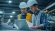© alauli - Two Asian engineers in hard hats and safety vests working together in a factory, reviewing documents on a laptop computer