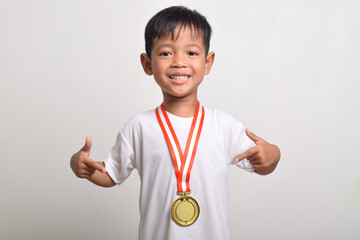 Asian boy with a finger showing his gold medal isolated on a white background. Smiling child celebrating his success. Trophy for the first place