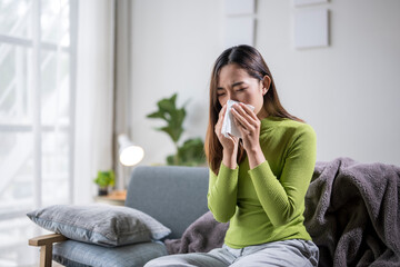 Sick asian woman sneezing in tissue at home on sofa