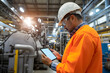 © Anastasia - A worker in an orange safety jacket and helmet uses a tablet in an industrial setting filled with machinery and equipment