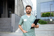 © zinkevych - Bearded doctor in his lab coat holding clipboard filled with notes standing outdoors hospital