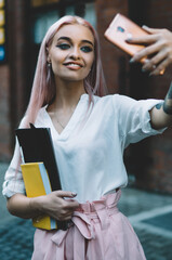 Happy female schooler with beautiful pink hair holding textbook for education and smiling while clicking selfie pictures via front camera on cellphone, cheerful millennial posing and taking images