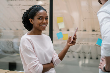 Portrait of happy cheerful afro american female financial director holding cellular phone in hand and smiling at camera while male colleague analyzing information from modern board during working time