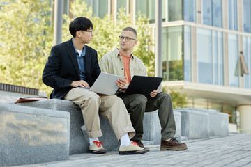 Full shot of male IT programmer with buzz cut in conversation with colleague using laptop to write code during outdoor meeting on bench in city street, copy space