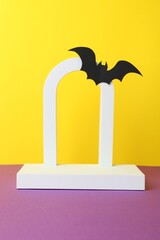 Canvas Print - Halloween celebration. Podium and paper bat on purple table against yellow background. Space for text