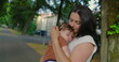 © Marco - Mother embracing her baby with a gentle smile on a tree-lined path, radiating warmth and affection in a serene urban setting with greenery and buildings in the background