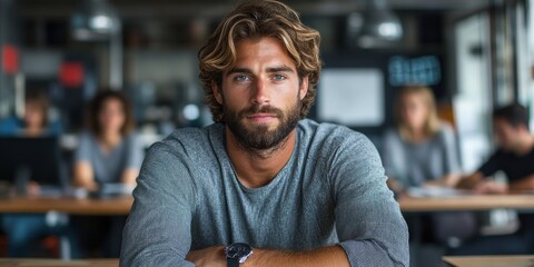 Wall Mural - A contemplative bearded man with wavy hair sits confidently at a modern office workspace with blurred colleagues in the background, representing a professional work environment