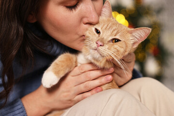 Wall Mural - Woman kissing her cute ginger cat against blurred Christmas lights indoors, closeup