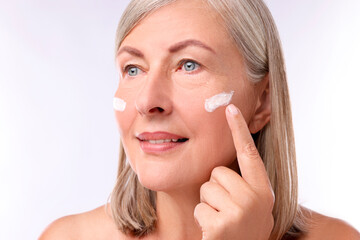 Poster - Senior woman applying face cream on white background