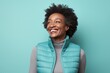 © Markus Schröder - Portrait of a joyful afro-american woman in her 40s dressed in a thermal insulation vest while standing against pastel teal background