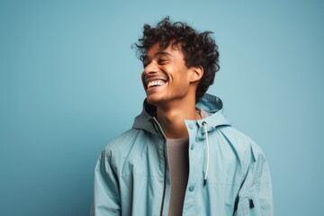 Portrait of a grinning indian man in his 20s wearing a functional windbreaker in front of soft blue background