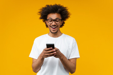 Studio shot of young smiling African American man in trendy eyeglasses reading message on his mobile phone with surprised face expression