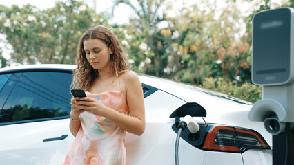 Young woman use smartphone to pay for electricity at public EV car charging station in nature. Modern environmental and sustainable automobile transportation lifestyle with EV vehicle. Synchronos