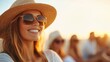 © Pinklife - A woman wearing a sun hat and sunglasses smiles joyfully at a beach during sunset, with friends blurred in the warm background, capturing a relaxed vacation vibe.