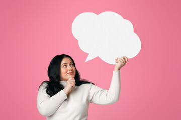 pensive plus size woman holding and looking at blank speech bubble over her head, standing on pink b