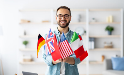 Modern education and student exchange. Cheerful young Caucasian guy holding bunch of different flags at home. Happy millennial man advertising foreign languages school, emigrating overseas