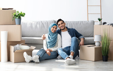 Cheerful young muslim family husband and wife in hijab posing while moving to their new apartment, sitting on floor by sofa among paper boxes with stuff, embracing and smiling at camera, copy space