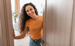© Prostock-studio - Portrait of cheerful young lady standing in doorway of new modern home, receiving and greeting visitor, happy smiling curly lady holding door looking out of slightly open ajar door