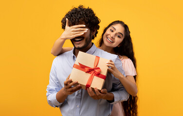 Pretty indian woman covering her boyfriend's eyes, holding wrapped gift box and greeting him with birthday or anniversary, standing over yellow studio background