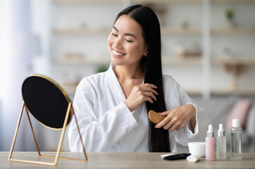 Wall Mural - Cheerful young asian lady in bathrobe sitting in bedroom, combing her beautiful silky hair with wooden brush, looking at mirror and smiling, copy space. Hair care products concept