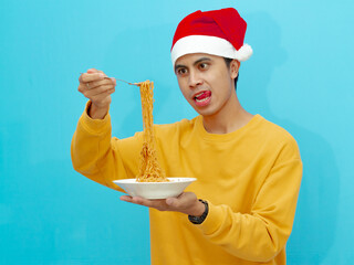 A hungry Asian man in a yellow sweater and Christmas hat picking fried noodles with a fork from a white plate, showing an appetizing expression. Isolated against a blue background.