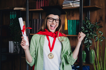 Photo of cheerful lucky schoolgirl dressed shirt mortarboard rising diploma indoors college auditorium