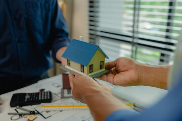 two architect is in office and sharing their strategy idea of building plan to each other, there are blueprint and house model on the desk in the office with two engineers, meeting