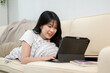 © bongkarn - A happy Asian woman is using her digital tablet while laying on a sofa in the living room.
