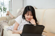 © bongkarn - A happy Asian woman is using her digital tablet while laying on a sofa in the living room.