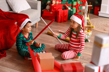 Cute little children in Santa hats wrapping Christmas gift at home