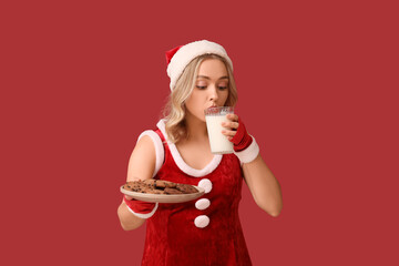 Wall Mural - Beautiful young woman dressed as Santa Claus drinking milk with tasty cookies on red background