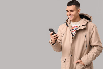 Wall Mural - Young man in stylish puffer jacket with mobile phone on grey background