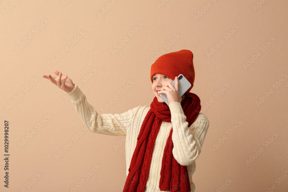 Cute little boy in winter clothes talking by mobile phone on beige background