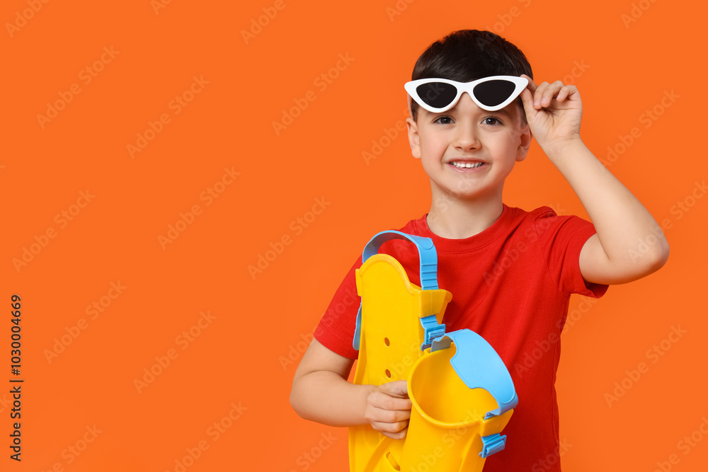 Cute little boy in sunglasses holding flippers on red background