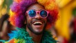 © Nataliia_Trushchenko - A person wearing a colorful clown wig and pink glasses, laughing heartily with their mouth open wide while standing against a yellow background. The individual is dressed in a green feather collar