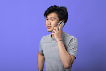 A young Asian man in a gray t-shirt is standing against a purple background, holding a smartphone to his ear as if engaged in a conversation. He has a casual stance with one hand on his hip