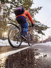 A man riding a bicycle on a bike path in winter in the city. Back view of a man in an orange jacket riding a bicycle. Ecological transportation in the city