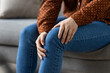 © Liubomir - Asian woman sitting on sofa holding knee, wearing jeans and polka dot top. Image captures concept of joint pain, health, and relaxation at home. Focus on discomfort and wellness.