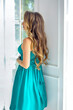 © Alina - Attractive beautiful brunette girl with curls and blue blowouts in a green midi dress. Light interior room with green flowers.