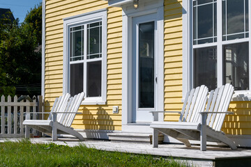 The front of a bright yellow wooden clapboard house. There's white trim, a white glass door and windows. Two double Adirondack chairs are on the sunny porch. There's a wood picket fence in the garden.