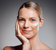 © KayExam/peopleimages.com - Portrait, happy woman and skincare cream for beauty, moisturizer and collagen. Lotion, mature model and apply cosmetics on face for dermatology, glow or shine for anti aging on gray studio background