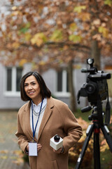 A confident multiracial journalist smiles with a microphone, prepared to report news in a peaceful autumn scene.